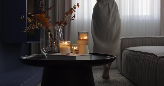 Unrecognizable young woman walk on tiptoes to the living room window. Modern living room interior design with blue TV cabinet sofa curtains and middle table with burning candles