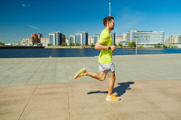 A man in a yellow shirt and shorts runs along a waterfront promenade on a sunny day, with city...