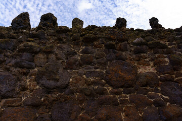 Old ruined battlement or crenellated wall, rough-textured wall constructed from irregularly shaped stones in shades of brown and black, ancient traditional Building technique, low-angle shot