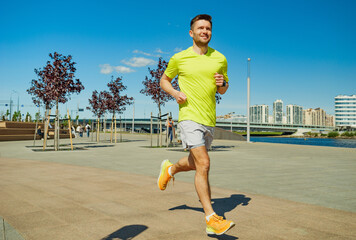 A man in a yellow shirt and shorts enjoys jogging in an urban park on a sunny day, with city...