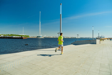 A man in a yellow shirt and shorts runs along a sunny waterfront promenade, with clear blue skies...