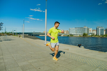 A man in a yellow shirt and shorts jogs along a sunny waterfront promenade, checking his...