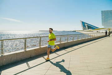 A man in a yellow shirt and shorts runs along a sunny waterfront promenade, with modern buildings...