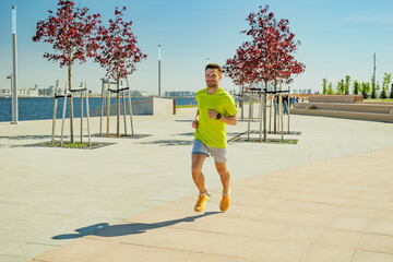 A man in a yellow shirt and shorts jogs through a sunny urban park, with red-leaved trees and a...