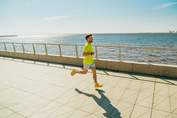A man in a yellow shirt and shorts jogs along a sunny waterfront promenade, with blue water and...