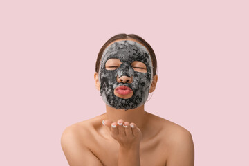 Young woman with activated charcoal mask on her face blowing kiss against pink background