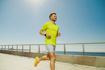 A man in a yellow shirt and shorts jogs along a sunny waterfront promenade, with the sun shining...
