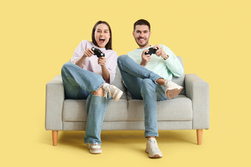 Beautiful young happy couple playing video games on sofa against yellow background