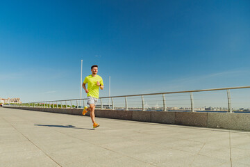 A man in a yellow shirt and shorts jogs along a sunny waterfront promenade, with clear blue skies...