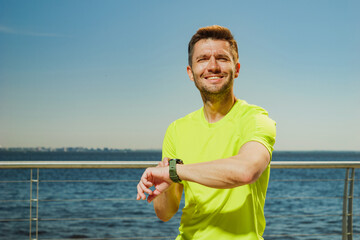 A man in a yellow shirt smiles while checking his wristwatch on a sunny waterfront promenade, with...