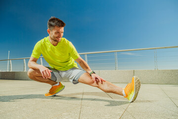 A man in a yellow shirt and shorts stretches outdoors on a sunny waterfront promenade, smiling as...