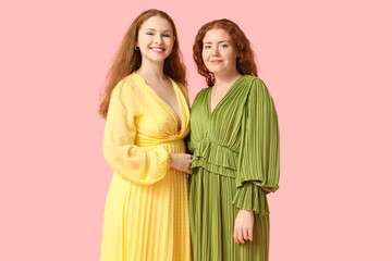 Happy redhead sisters in stylish dresses on pink background