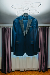 A blue tuxedo jacket hanging from a ceiling fixture in a room. The jacket is neatly arranged with its lapels facing outward. The room has a window with purple curtains