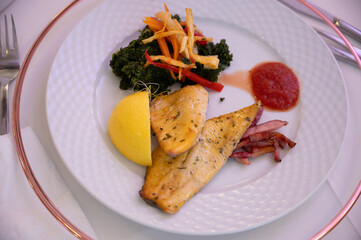 A plate of food placed on a table. The plate contains a piece of grilled fish, a slice of lemon, a portion of sautéed vegetables, and a small dab of red sauce. The plate is white