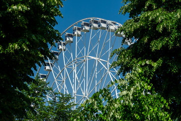 A large ferris wheel set against a clear blue sky. The wheel is constructed with white metal beams and has multiple cabins attached to its circumference