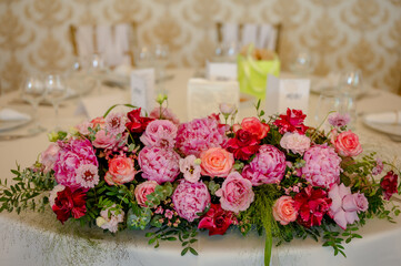 A beautifully set dining table adorned with a vibrant floral centerpiece. The centerpiece is dominated by pink and red flowers, possibly roses, interspersed with green foliage.