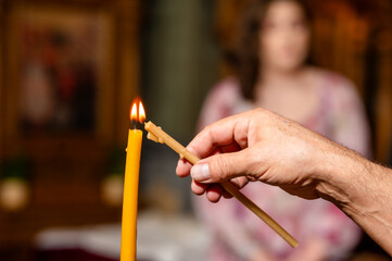 A close-up of a hand lighting a candle. The candle is tall, slender, and of a bright yellow color. The hand holding the candle's wick is slightly blurred, emphasizing the act of lighting