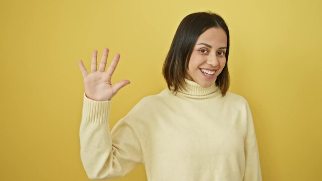 Cheerful, confident young hispanic woman in sweater strikes a pose, happily showing off a high five gesture, all smiles on an isolated yellow background