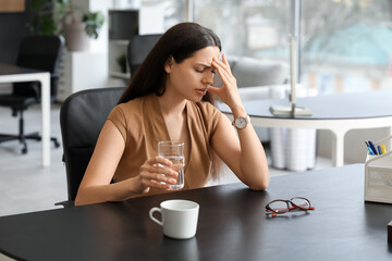 Young woman with glass of water suffering from headache sitting in office