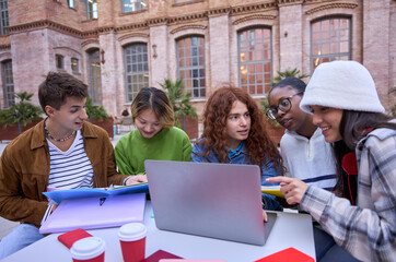 Group of smiling and concentrated diverse students using laptop and talking doing brainstorming on university campus. Young people friends sitting working and studying together outdoor