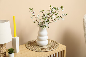 Blooming branches in vase, wicker mat and candle on table near beige wall