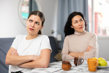 Asian and European offended women sitting at table at home.