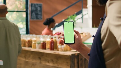Local seller holds smartphone app with greenscreen display, presenting copyspace technology on mobile device. Young man showing isolated mockup screen at grocery store checkout.