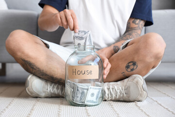 Young man putting money into jar for house at home, closeup