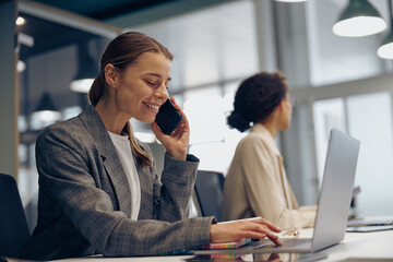 Woman sales manager talking by phone with client sitting in modern coworking and working on laptop
