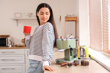 Beautiful young woman with modern coffee machine and cup of cappuccino in kitchen