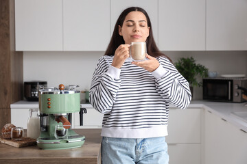 Beautiful young happy woman with modern coffee machine and cup of latte in kitchen