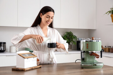 Beautiful young happy woman with modern machine pouring milk into cup of fresh coffee in kitchen