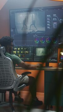 African American male editor makes color grading, works at home office. Action movie footage and program interface on multi-monitor computer and big digital screen. Vertical shot