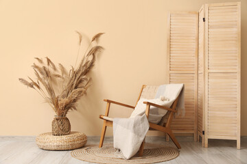 Comfortable armchair with wicker pouf and folding screen near beige wall