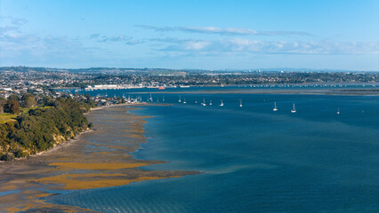 Aerial view of a coastal town with numerous sailboats anchored in a calm blue bay, under a clear sky - Musick Point, Auckland