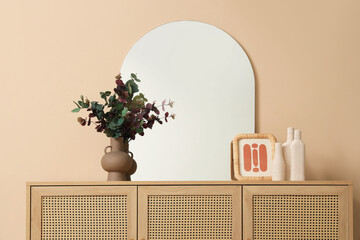 Flowers in vase, mirror, bottles and frame on wooden chest of drawers near beige wall