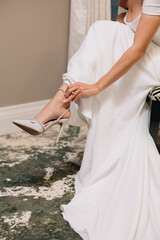 The bride in a white wedding dress puts on high heeled shoes. The bride is preparing for the wedding ceremony. Beautiful sparkling high heel wedding shoes close up