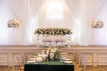 A large festive wedding hall. The wedding table is decorated with white flowers and two white chairs for the newlyweds stand on the stage. The hall is decorated for a wedding and a festive table