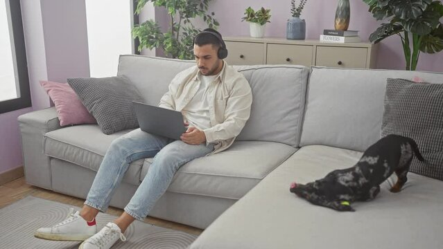 A young hispanic man works on his laptop at home while his teckel dog plays on the couch.