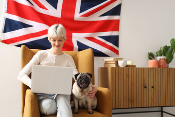 Young woman with pug dog sitting in armchair and hanging UK flag on wall at home