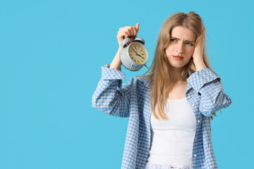 Young woman in pajamas with alarm clock suffering from insomnia on blue background
