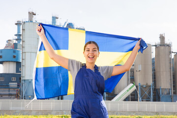 Cheerful young woman in workwear holds the national flag of Sweden in her hands, standing on the...