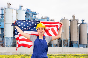 Positive young female worker in helmet waving national flag of USA while standing in front of big...