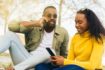 afro american couple sitting on the grass using mobile and talking on the phone