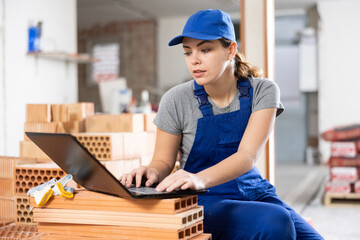 Focused young female contractor in blue uniform using laptop to check project while working at...