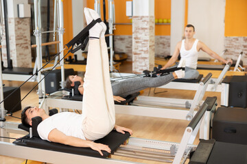 Male gym visitor performs exercises to increase flexibility and stretch leg muscles on reformer....