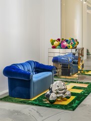In one corner of the gallery stands a colorful sideboard with a sculpture of stacked stuffed teddy bears in different colors, a beige green patterned carpet covers the floor, and blue velvet sofas lea