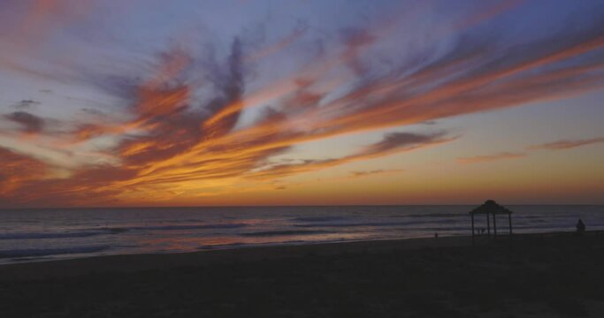 People silhouettes walking on beach next to ocean wavew at calm sunset with purple and orange clouds