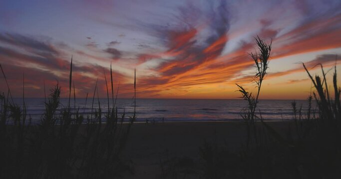 Beach at sunset with dramatic orange and purple sky. People walking on sand next to ocean with sur boards at dusk.