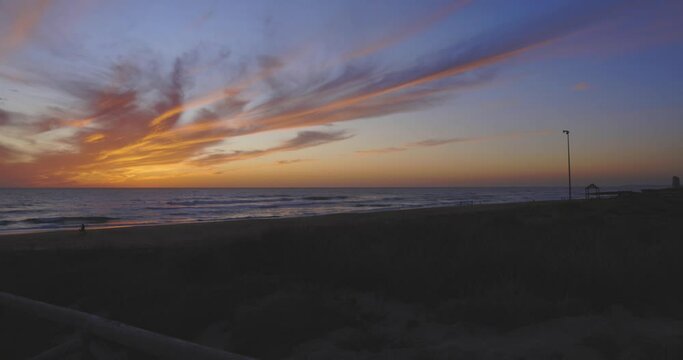  People walking on beach at sunset with dramatic ocean orange and purple sky next to gazebo with walkway.